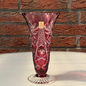 Könnte beinhalten: Eine rubinrote Kristallvase mit ausgestelltem Oberteil und detailliertem Schliffdesign. Die Vase hat einen runden Fuß mit Sternmuster und ein kleines Etikett in der Nähe der Oberseite. Die Vase ist etwa 25 cm hoch.