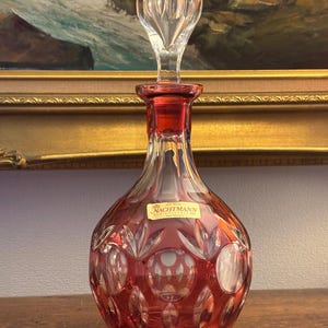 Può includere: Una caraffa in vetro trasparente Nachtmann con una tonalità rossa e un tappo trasparente. La caraffa ha un corpo bulboso con dettagli in vetro tagliato. Il tappo ha un design trasparente e sfaccettato. La caraffa è su una superficie di legno.