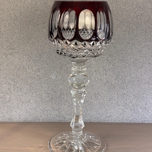 May include: A tall, elegant wine glass with a deep red bowl and a clear, intricately cut stem and base. The bowl features vertical oval cuts and diamond-shaped patterns. The glass stands on a round base with a starburst design.