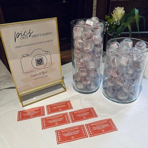 Könnte beinhalten: Eine Tischdekoration mit einem goldgerahmten Schild mit der Aufschrift "pics or it didn't happen" und einer Kamera-Grafik. Zwei Glasvasen sind mit transparenten Kugeln mit roten Akzenten gefüllt. Rote "Photo Challenge"-Karten liegen verstreut.