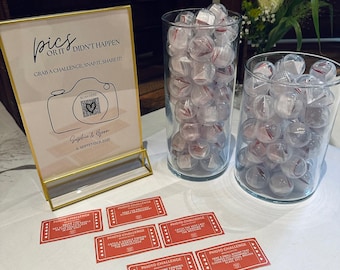 May include: A table setting with a gold-framed sign that reads "pics or it didn't happen" and a camera graphic. Two glass vases are filled with clear, red-accented spheres. Red "Photo Challenge" cards are scattered on the white tablecloth.