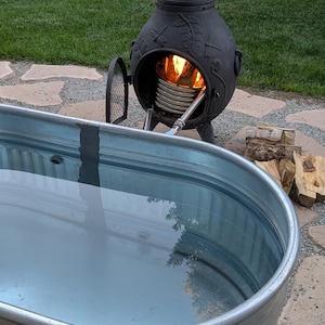 May include: A galvanized metal stock tank filled with water, next to a black outdoor heater with a fire burning inside. Firewood is stacked nearby on a stone patio, with green grass in the background. The scene suggests outdoor relaxation.
