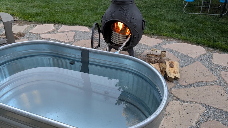 May include: A galvanized metal stock tank filled with water, next to a black outdoor heater with a fire burning inside. Firewood is stacked nearby on a stone patio, with green grass in the background. The scene suggests outdoor relaxation.