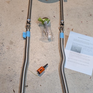 May include: A stainless steel immersion chiller coil with two flexible hoses, valves, and fittings. Additional components include a green-handled tool, an orange hole saw, and a printed instruction sheet. The items are arranged on a concrete surface.