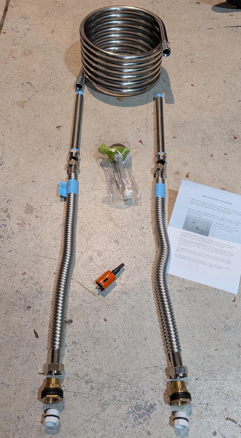 May include: A stainless steel immersion chiller coil with two flexible hoses, valves, and fittings. Additional components include a green-handled tool, an orange hole saw, and a printed instruction sheet. The items are arranged on a concrete surface.