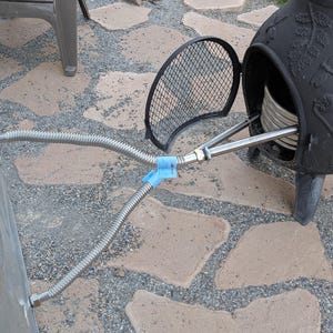 May include: Outdoor setup featuring a galvanized metal tub connected to a black metal chiminea with a hinged door. Flexible metal hoses and a blue plastic clip are visible. The chiminea has decorative floral patterns.