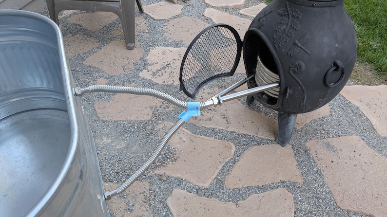 May include: Outdoor setup featuring a galvanized metal tub connected to a black metal chiminea with a hinged door. Flexible metal hoses and a blue plastic clip are visible. The chiminea has decorative floral patterns.
