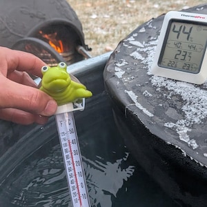 May include: A green frog-shaped thermometer is placed in water, showing a temperature reading. A digital thermometer displays the temperature in Fahrenheit and Celsius. The background includes a fire pit and outdoor seating.