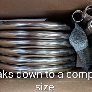 May include: A collection of shiny, silver metal coils and fittings, neatly arranged inside a cardboard box. The text "Breaks down to a compact size" is visible. The components appear to be plumbing or industrial parts.