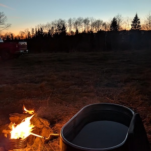 May include: A campfire with flames and a metal tub filled with water. The scene is set outdoors at dusk, with a dark treeline and a colorful sky in the background. The tub is near the fire.