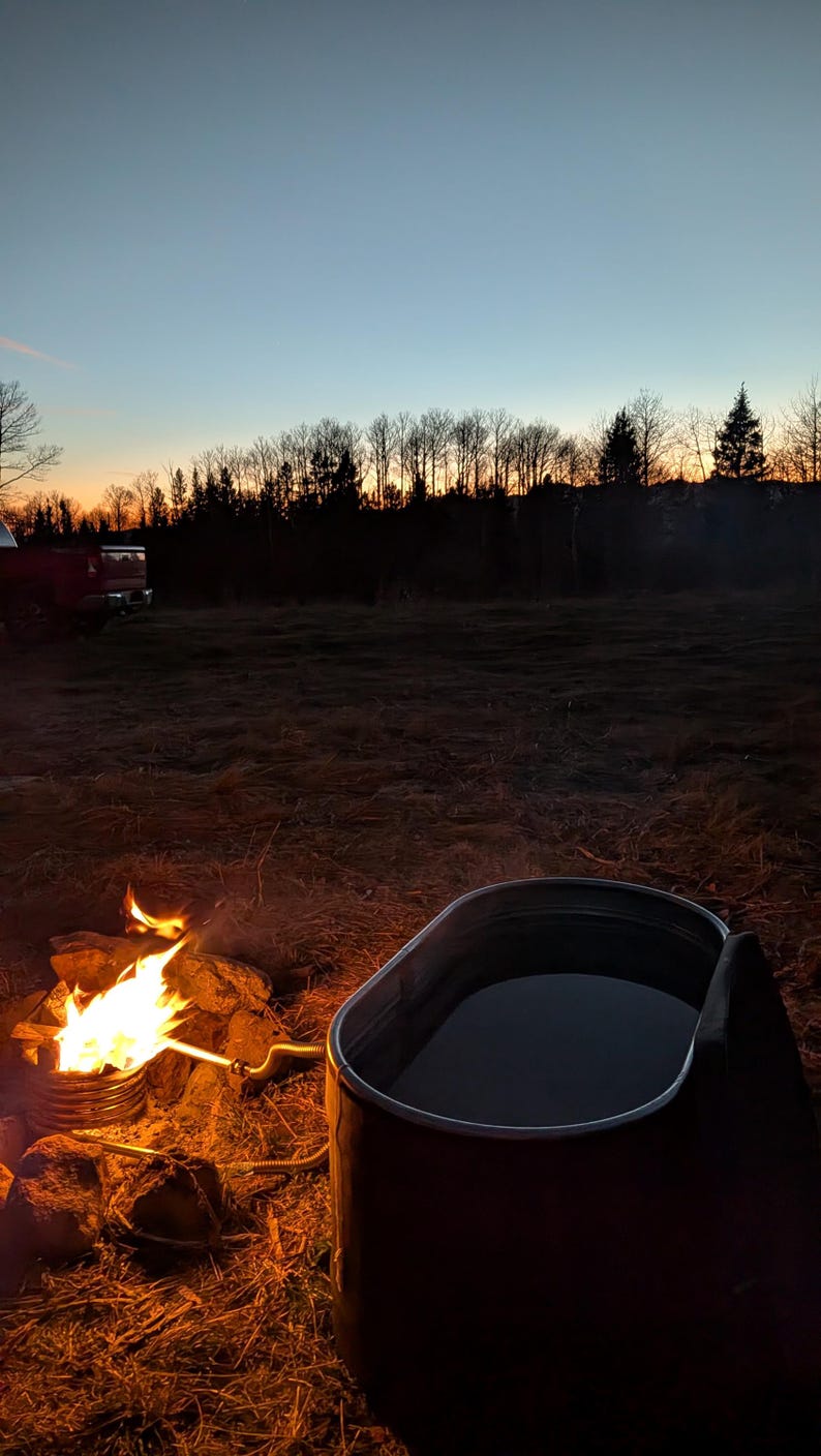 May include: A campfire with flames and a metal tub filled with water. The scene is set outdoors at dusk, with a dark treeline and a colorful sky in the background. The tub is near the fire.