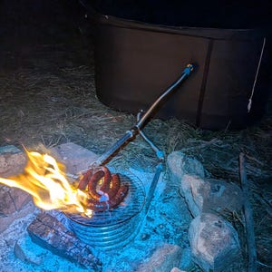 May include: A campfire scene at night with flames, a grill, and sausages. The grill is over a bed of glowing embers and surrounded by rocks. A metal arm extends over the grill. A dark tent is in the background.