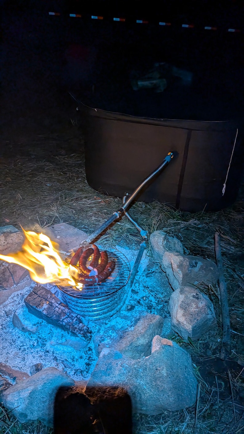 May include: A campfire scene at night with flames, a grill, and sausages. The grill is over a bed of glowing embers and surrounded by rocks. A metal arm extends over the grill. A dark tent is in the background.