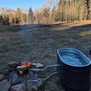 May include: An outdoor setup featuring a black metal stock tank filled with water, connected by pipes to a fire-heated coil. A small fire burns with firewood, surrounded by rocks, in a natural outdoor setting with trees.