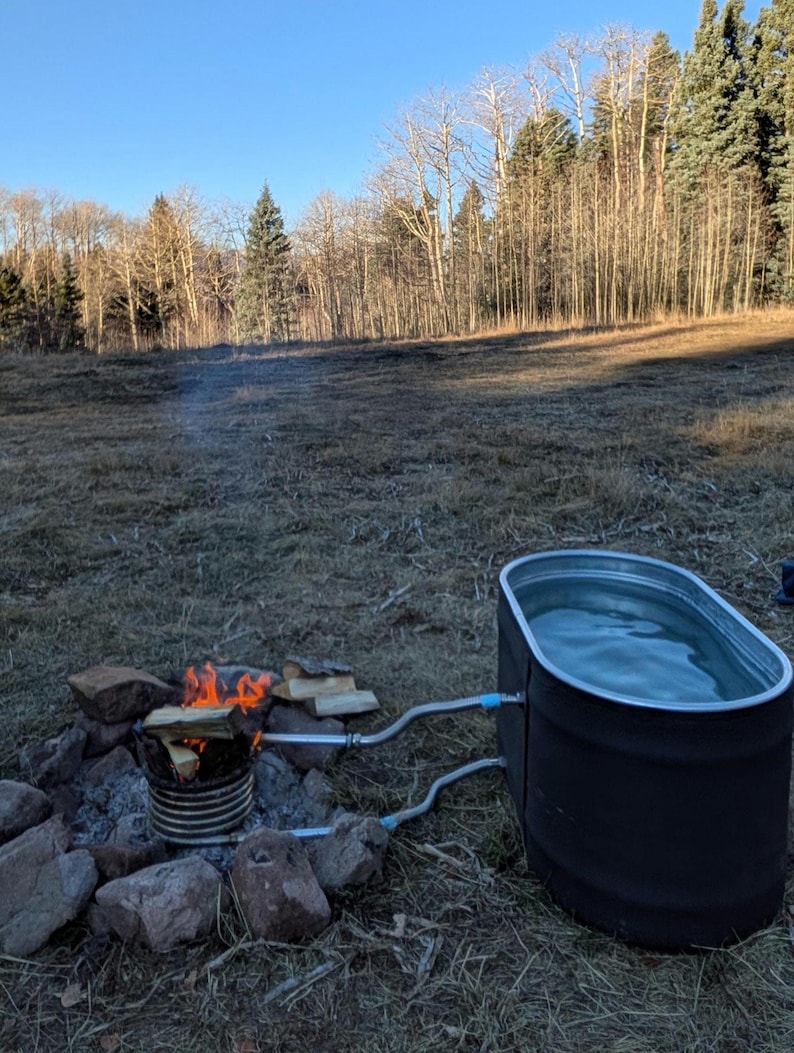 May include: An outdoor setup featuring a black metal stock tank filled with water, connected by pipes to a fire-heated coil. A small fire burns with firewood, surrounded by rocks, in a natural outdoor setting with trees.