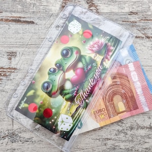 May include: A clear plastic binder pouch with a colorful illustration of two cartoon frogs embracing, holding a heart, with the words "Thank you". Euro banknotes are visible inside the pouch.