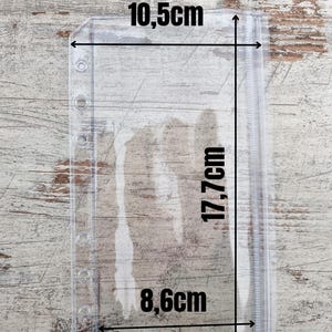 May include: Clear plastic A6 binder pocket with measurements: 10.5cm wide, 17.7cm tall, and 8.6cm wide. The text "Passend f&uuml;r den A6 Binder" is at the top. The pocket has holes for binding and a zipper closure.