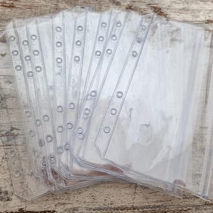 May include: A stack of clear, transparent plastic binder sleeves. Each sleeve has a row of holes along the left edge for insertion into a binder. The sleeves are rectangular with a sealed bottom and open top for inserting documents or other items.