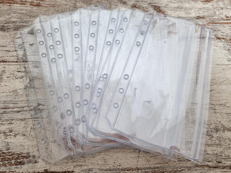 May include: A stack of clear, transparent plastic binder sleeves. Each sleeve has a row of holes along the left edge for insertion into a binder. The sleeves are rectangular with a sealed bottom and open top for inserting documents or other items.