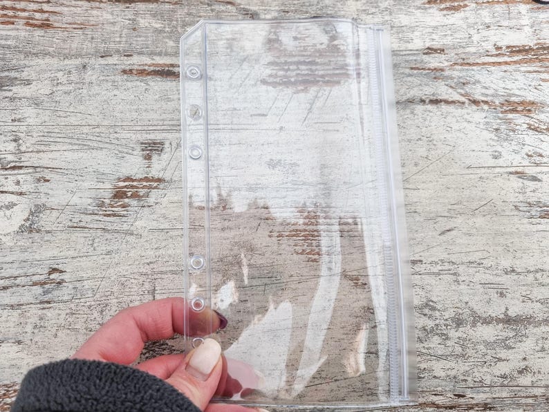 May include: A clear, plastic binder sleeve with a zipper closure and several holes along the left edge for insertion into a binder. The sleeve is held against a weathered, light-colored wooden surface.