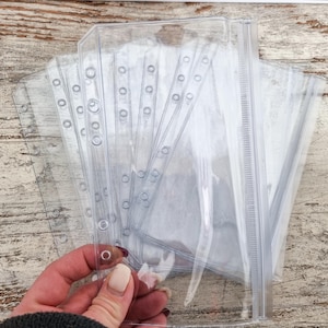 Könnte beinhalten: Transparente Kunststoff-Ordner-Taschen mit verstärkten Kanten und Löchern zum Einlegen in einen Ordner. Die Taschen haben einen Reißverschluss und sind für die Aufnahme von Dokumenten oder anderen Gegenständen konzipiert.