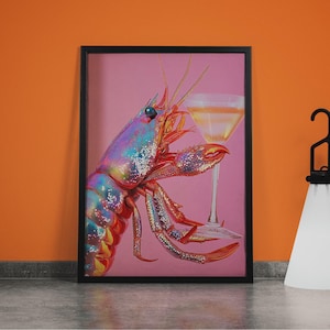 Op de afbeelding: Een ingelijste kunstdruk met een kleurrijke kreeft die een martini glas vasthoudt. De kreeft heeft iriserende blauwe, paarse en rode tinten. De achtergrond is roze en de lijst is zwart. Het martini glas bevat een gele vloeistof.