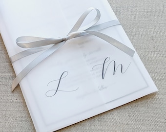 May include: A white wedding invitation with a silver ribbon tied in a bow. The invitation has the initials "L" and "M" written in calligraphy.