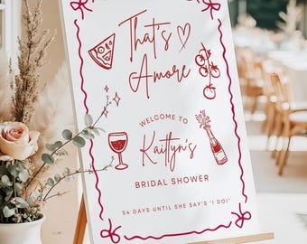 May include: A white sign on a wooden easel with the text "That's Amore" and "Welcome to Kaitlyn's Bridal Shower" in burgundy script. The sign includes illustrations of pizza, wine, and champagne.