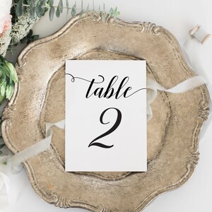May include: A white card with black calligraphy that reads "Table 2" on a silver charger with a white ribbon.