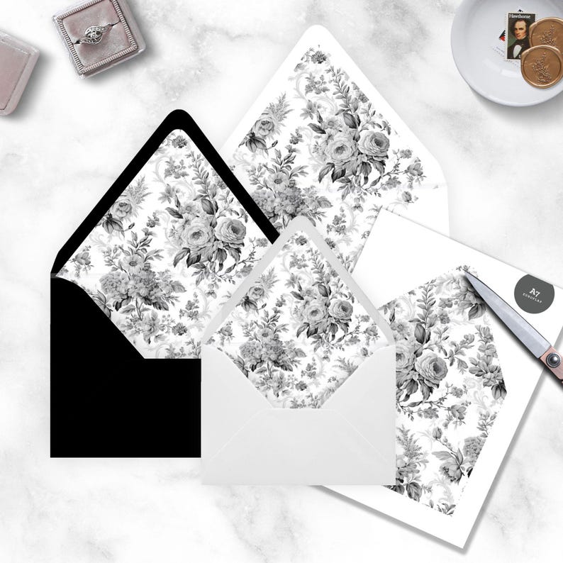 May include: Three white envelopes with a black and white floral pattern. The envelopes are nested inside each other, with the largest envelope at the bottom and the smallest envelope at the top. The envelopes are on a white marble surface.