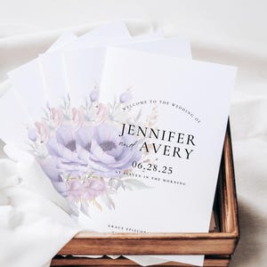 May include: A collection of white wedding programs featuring purple and pink floral illustrations, presented in a wooden tray. The programs display the text "Welcome to the Wedding of Jennifer and Avery 06.28.25".