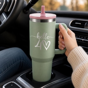 Könnte beinhalten: Ein salbeigrüner Edelstahlbecher mit Henkel, rosa Deckel und passendem Trinkhalm. Auf der Vorderseite des Bechers ist der weiße Schriftzug hello und die Zahl 40, gefolgt von einem Herzsymbol, in einer geschwungenen Schriftart eingraviert.