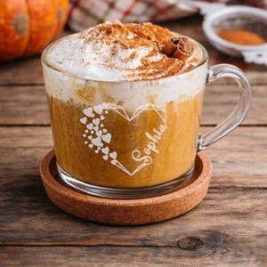 Op de afbeelding: Een witte keramische mok met een zwarte handgreep en de tekst "But First, Coffee" in een vetgedrukte, zwarte letter. De mok staat tegen een vage achtergrond. De mok is ongeveer 10 cm hoog.