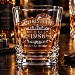 Könnte beinhalten: Ein klares Whiskyglas gefüllt mit bernsteinfarbener Flüssigkeit und Eis, mit einer Gravur, die CHRISTIAN LIMITED EDITION 1986 PREMIUM JAHRGANG zeigt. Das Glas hat einen schweren, facettierten Boden und glatte Seiten, die den Text hervorheben.