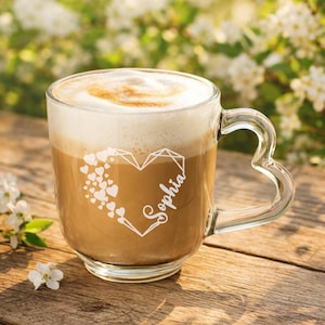 Op de afbeelding: Een heldere glazen koffiemok met een hartvormig handvat, gevuld met een latte en bedekt met schuim. De mok heeft een hartontwerp met de naam "Sophia" in script. De achtergrond is wazig met groen en zonlicht.