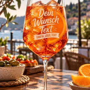 May include: A clear stemmed glass filled with an orange cocktail, ice cubes, and an orange slice. The glass features white text that reads "Dein Wunsch Text" above a banner with the words "GRAFIK, LOGO, TEXT". A black straw is inserted into the drink.