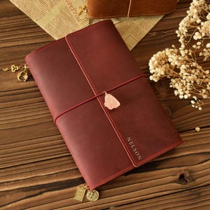 May include: A burgundy leather journal with a red elastic closure and a rose quartz charm. The journal has the name "NELSON" embossed on the cover and is adorned with gold-colored charms. The journal is lying on a wooden surface.