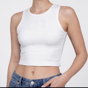 May include: A white ribbed tank top with a crew neck and a cropped length. The top is sleeveless and has a fitted silhouette. The model is wearing blue jeans. The overall style is casual and minimalist.