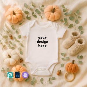 May include: A white baby onesie with the text "your design here" is surrounded by fall-themed decorations. The decorations include small pumpkins, knitted baby booties, acorns, and eucalyptus leaves. A wooden teething ring is also present.