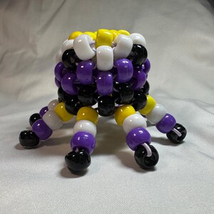 May include: A handmade, beaded octopus sculpture in non-binary pride colors. The body is a cube of white, yellow, purple, and black beads. Eight beaded tentacles extend from the body, each with a black bead at the end. The sculpture is on a white background.