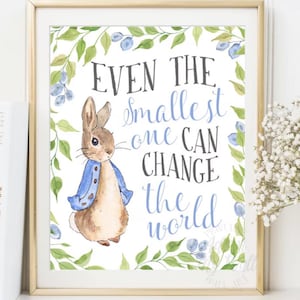 May include: Framed art print featuring a watercolor illustration of Peter Rabbit wearing a blue jacket. The print is surrounded by green leaves and blueberries, with the text "Even the smallest one can change the world" in black and blue.