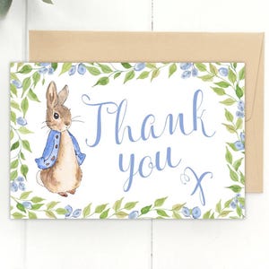 May include: A watercolor thank you card featuring Peter Rabbit in a blue jacket, surrounded by a green and blue floral border. The words "Thank you" are written in light blue script, with a brown envelope behind the card.