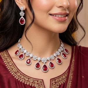 May include: A stunning necklace and earring set. The necklace and earrings feature teardrop-shaped, deep red gemstones, each encircled by sparkling white stones. The set is worn with a maroon garment.