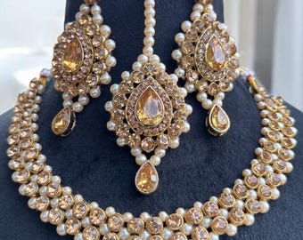 Pode incluir: Um conjunto de joias douradas com colar, brincos e um adorno de cabeça. O colar e os brincos apresentam um design em forma de gota com pedras âmbar e pérolas falsas. O adorno de cabeça tem uma pedra âmbar central e uma fileira de pérolas.