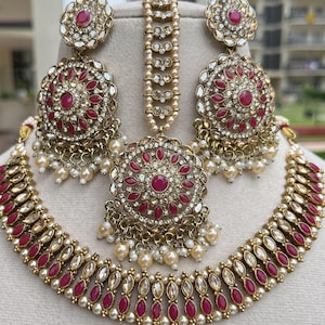 May include: A gold-toned jewelry set featuring a necklace, earrings, and a headpiece. The set includes ruby-colored stones and small white pearls. The earrings and necklace have a circular design with intricate detailing.