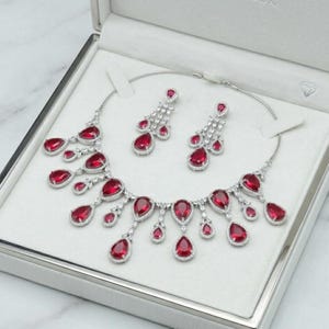 May include: A jewellery set comprising a necklace and matching earrings, displayed in a white box. The set features a silver-toned necklace and earrings embellished with teardrop-shaped, deep red gemstones, framed by smaller, clear stones.