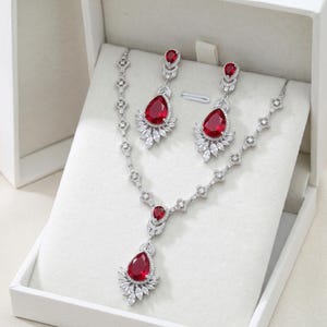 May include: A jewelry set featuring a necklace and earrings with a silver-tone metal and red teardrop-shaped stones. The earrings and necklace have a cluster of white stones around the red stones. The set is displayed in a white box.
