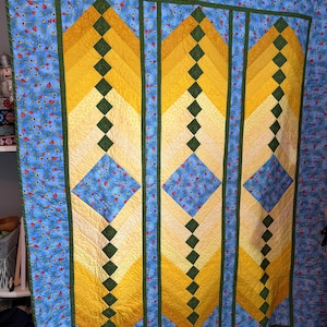 May include: A quilt featuring a blue fabric with a red and yellow floral pattern, accented by vertical panels of yellow and cream ombre fabric arranged in a chevron design, separated by thin dark green strips and small green squares.