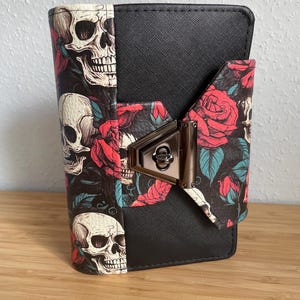 Könnte beinhalten: Eine schwarze Geldbörse mit strukturierter Oberfläche und einem Frontpanel, das mit einem Muster aus weißen Totenköpfen, roten Rosen und blaugrünen Blättern verziert ist. Die Geldbörse wird mit einem metallischen Drehverschluss gesichert.