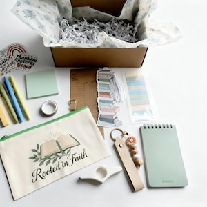 May include: A stationery gift set featuring a beige zippered pouch with a book graphic and the text Rooted in Faith, a sage green spiral notebook, pens, sticky notes, a book-shaped bookmark, a keychain, and stickers, all packed in a brown box.